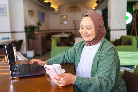 A Muslim woman in a hijab is seated at a cafe table with a laptop, holding a phone, and smiling.