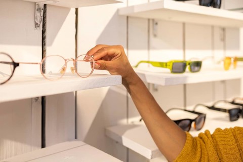 A hand reaches out to select a stylish pair of glasses from a minimalist white display shelf in a