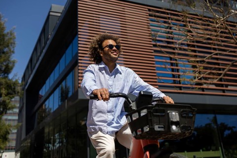 A relaxed man with sunglasses rides a public bicycle outside a modern building, enjoying a break