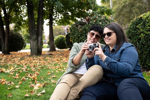 A lesbian curvy couple enjoys an autumn day seated on grass, capturing memories with a camera. The