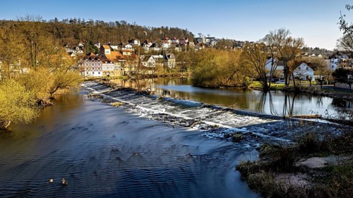 Small waterfall in a river in front of a village with autumnal trees and a clear sky, The weir of