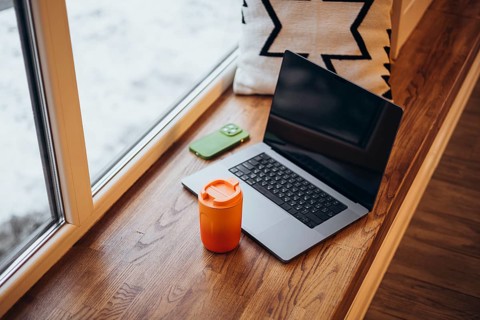 A cozy remote work setup featuring a laptop and a bright orange thermos cup beside a snowy window,