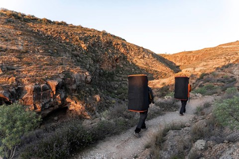 Two climbers carry crash pads along a rocky path surrounded by stunning natural scenery, preparing