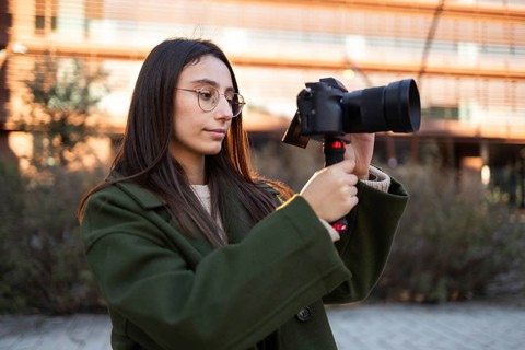 A young woman wears a green coat, carefully using a camera outdoors With clear glasses and long
