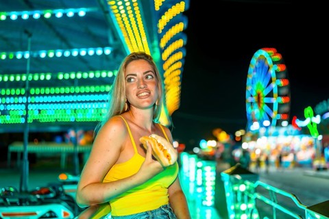Blonde woman holding an unhealthy hot dog in a night fair in summer