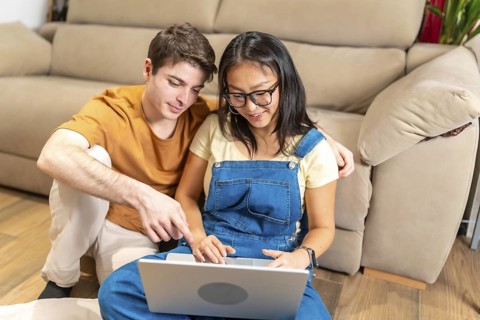 Young multiethnic couple sitting comfortably on the floor of their cozy living room, using a laptop