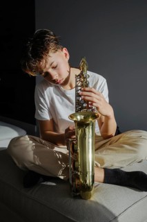 A young boy sits on a couch, holding a saxophone, bathed in soft natural light. His expression is