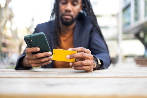 A cuban businessman sits at a wooden table outdoors, holding a smartphone and a yellow card,