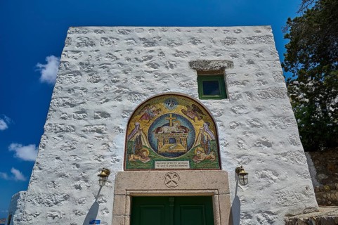 A white building with a green door and an ornate mosaic on a stone wall under a clear sky,