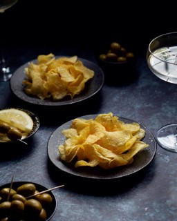 A close-up of golden potato chips on rustic black plates. Accompanied by green olives and sliced