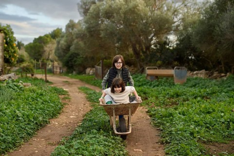 Two children enjoy a playful moment as one pushes the other in a wheelbarrow down a grassy, rural