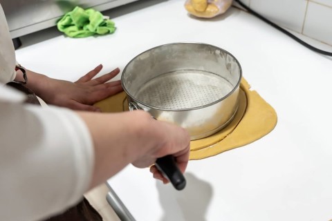 Baker hands preparing shortcrust pastry by cutting a circular dough base to fit a metal springform