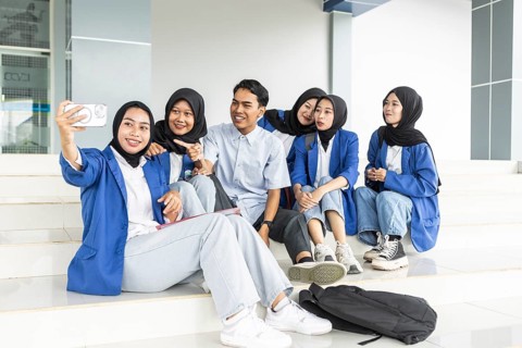 A diverse group of students, including Asian Muslim women, take a selfie on the steps of a school