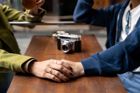 A close-up of a lesbian couple holding hands across a wooden table. A vintage camera and a glass of