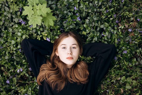 A teenage girl lies peacefully on a bed of lush periwinkle flowers in a tranquil forest setting,