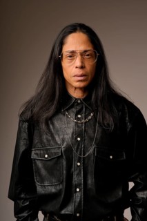 Close-up portrait of a Latin man wearing a black leather shirt and glasses, posing confidently. The