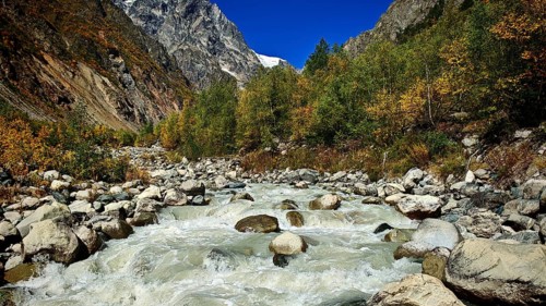 Stormy river flows through a valley surrounded by forested mountains under blue skies, hiking to