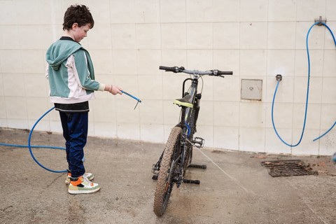 A young boy cleans a muddy mountain bike with a blue hose against a tiled wall. The scene captures