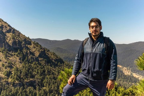A man with a backpack stands confidently on a mountain trail, surrounded by lush, forested hills