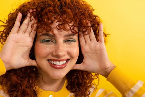 A smiling woman with curly red hair poses playfully against a bright yellow background,