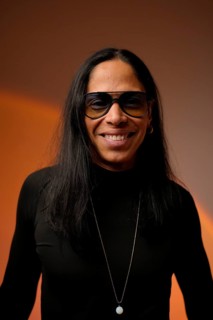 Latin man with long hair, wearing glasses and a necklace, smiles warmly. The studio background is