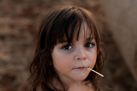 A thoughtful child with long hair gazes into the camera, holding a stick in their mouth. The soft,