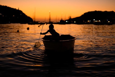 A silhouette of a child in a small boat with a net, gently floating on calm waters during a