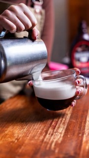 A barista artfully pours frothy milk into a glass espresso cup, creating a rich coffee blend. The