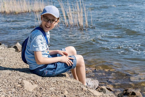 A joyful boy sits on a rocky lakeside, wearing casual summer attire and a cap, with a backpack,