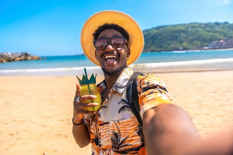 Young man savoring a tropical cocktail on a sunny beach, capturing the moment with a selfie while