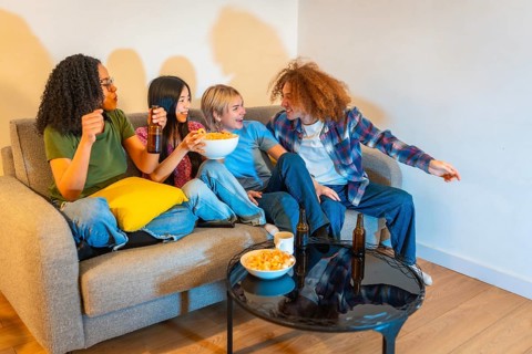 Four young friends enjoying their free time watching television, eating snacks and drinking beer,