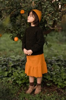 A young girl in a cozy knit sweater and orange skirt gazes at oranges on a tree. She stands in a