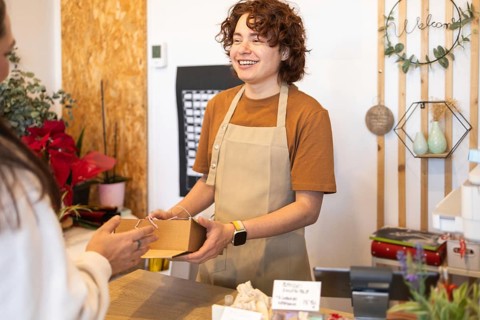 A smiling person in a shop gives a gift box to a customer The decor includes plants and wooden
