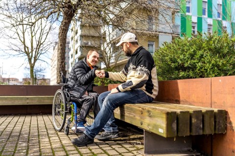 Young man in a wheelchair shakes hands with a friend sitting next to him on a bench in a city park