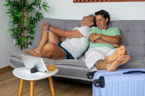 A joyful lesbian couple lounges on a sofa, laughing, with a suitcase nearby as they plan their