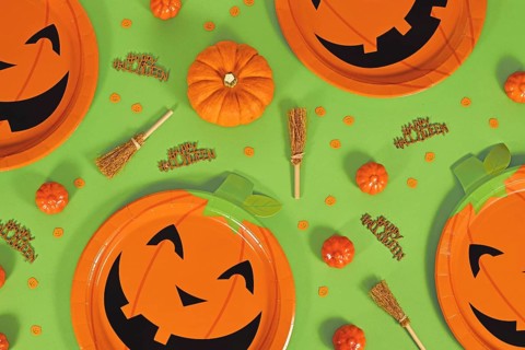 Halloween party flat lay with orange pumpkin paper plates, witch brooms, confetti and Happy