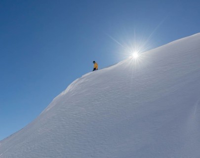 A snowboarder in a yellow jacket glides down a pristine snowy slope with the sun shining brightly