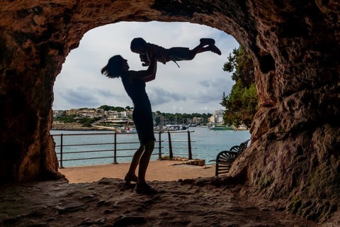 A woman is holding a child in her arms while standing in front of a large rock. The scene is