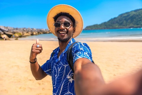 Young black man with straw hat and sunglasses gesturing thumbs up while taking a selfie on a sunny