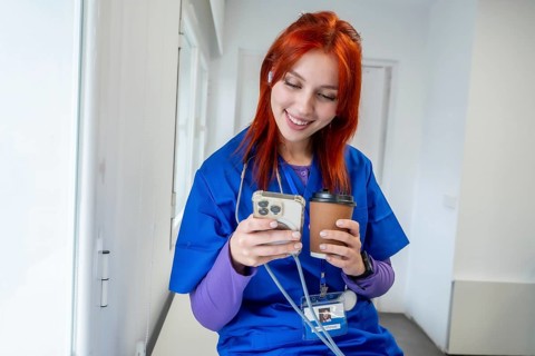 Young nurse enjoying a coffee break while scrolling on smartphone, wearing scrubs and an earbud,