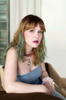 A woman with blonde and teal hair poses indoors wearing pastel pink makeup and a blue top She has a