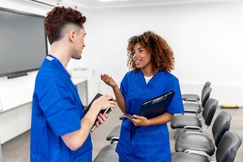 Two diverse young medical students wearing blue scrubs conversing in a modern university classroom,