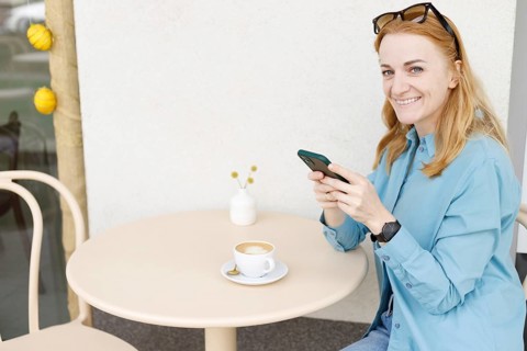 Smiling woman sitting at an outdoor cafe in Warsaw, using her smartphone and enjoying a cup of