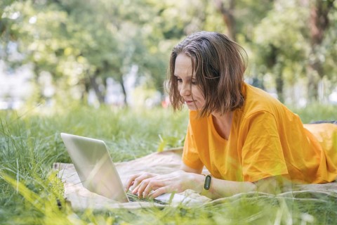 A woman lies on a blanket in a grassy park, focused on a laptop. She enjoys the outdoors while