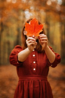 A woman in a rustic red dress holds a vibrant orange maple leaf in a forest, capturing the essence