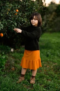 A young child dressed in a cozy dark sweater and orange skirt is picking oranges from a lush green