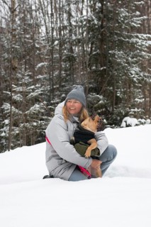 Woman in a winter coat and beanie happily embracing a dog in a snowy forest setting. The serene