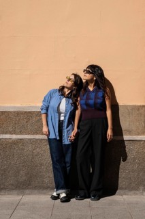 A lesbian couple stands outdoors, holding hands and basking in the sun. They celebrate love and