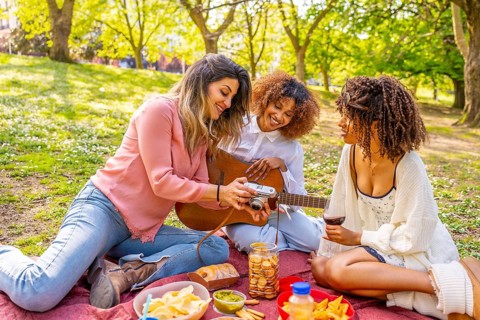 Three women relaxing on blanket, sharing food, playing guitar, and taking photos during sunny