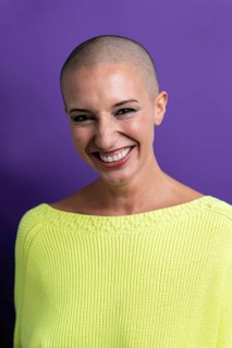 A middle-aged woman with a shaved head smiles warmly, wearing a neon green sweater against a